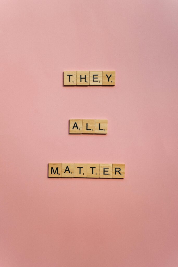 Scrabble tiles spelling 'They All Matter' on a soft pink background.